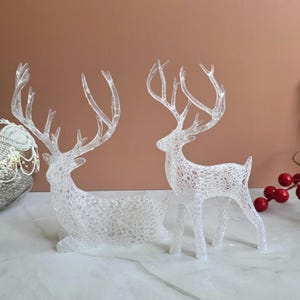 Puede incluir: Dos figuras decorativas transparentes de renos. Uno está tumbado, el otro de pie. Ambos tienen cuernos detallados y un diseño calado. Se colocan sobre una superficie blanca con un fondo marrón, lo que sugiere un tema navideño o invernal.