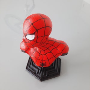 May include: A red Spider-Man bust figurine with a black web pattern and white eye details. The bust is mounted on a black geometric base, showcasing a detailed representation of the superhero's head and shoulders.