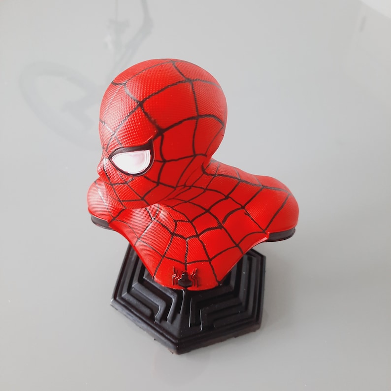 May include: A red Spider-Man bust figurine with a black web pattern and white eye details. The bust is mounted on a black geometric base, showcasing a detailed representation of the superhero's head and shoulders.