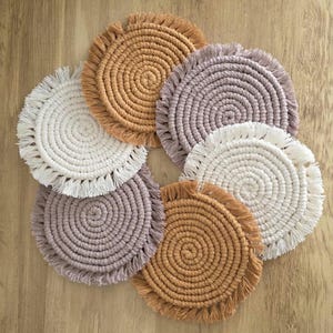 Puede incluir: Un conjunto de siete posavasos de macramé hechos a mano en varios colores, incluyendo blanco, marrón y lavanda. Cada posavasos presenta un diseño en espiral y un borde con flecos. Los posavasos están dispuestos sobre una superficie de madera.