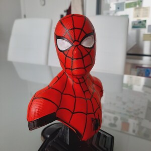 May include: A red and black Spider-Man bust with white eye lenses. The bust features a detailed web pattern and a black base. The superhero figure is a collectible item.