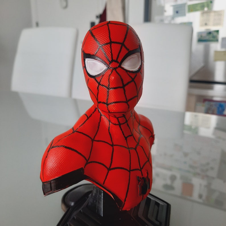 May include: A red and black Spider-Man bust with white eye lenses. The bust features a detailed web pattern and a black base. The superhero figure is a collectible item.