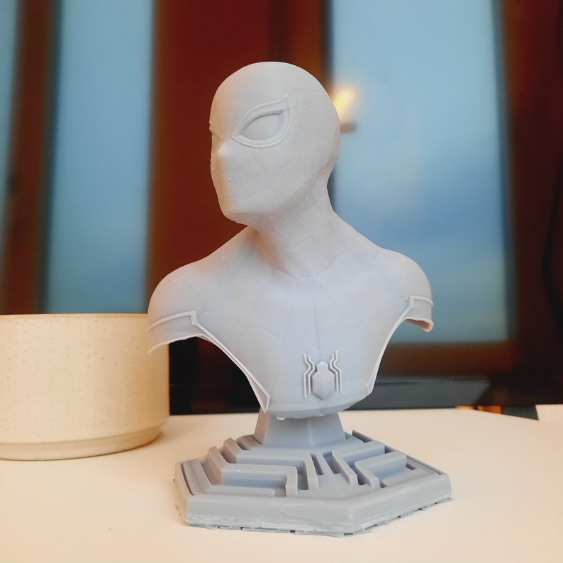 May include: A light gray bust of Spider-Man, featuring the character's mask and chest emblem. The bust is set on a geometric base and is likely a collectible item or decorative piece. The bust is made of a light gray material.
