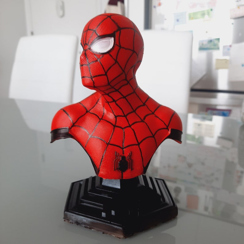 May include: A red and black Spider-Man bust figurine. The bust features a detailed depiction of Spider-Man's mask and costume, with a black spider emblem on the chest. The figurine is mounted on a black geometric base.