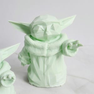 Pu&ograve; includere: Statuetta geometrica verde chiaro di un personaggio con grandi orecchie e una veste. La statuetta &egrave; in posizione eretta con un braccio teso. La figura &egrave; di colore verde chiaro e ha una superficie strutturata.