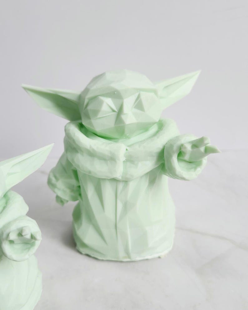 Pu&ograve; includere: Statuetta geometrica verde chiaro di un personaggio con grandi orecchie e una veste. La statuetta &egrave; in posizione eretta con un braccio teso. La figura &egrave; di colore verde chiaro e ha una superficie strutturata.