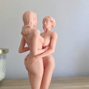May include: A nude sculpture of two figures embracing. The figures are in a light peach color, with detailed facial features and hair. The sculpture is a representation of human form and connection.