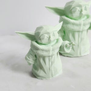 Pu&ograve; includere: Due sculture geometriche verde chiaro di un personaggio con grandi orecchie e un mantello con cappuccio. Le sculture hanno un design sfaccettato, che conferisce loro un aspetto moderno e angolare. Una scultura &egrave; in primo piano, con un'altra leggermente sfocata sullo sfondo.
