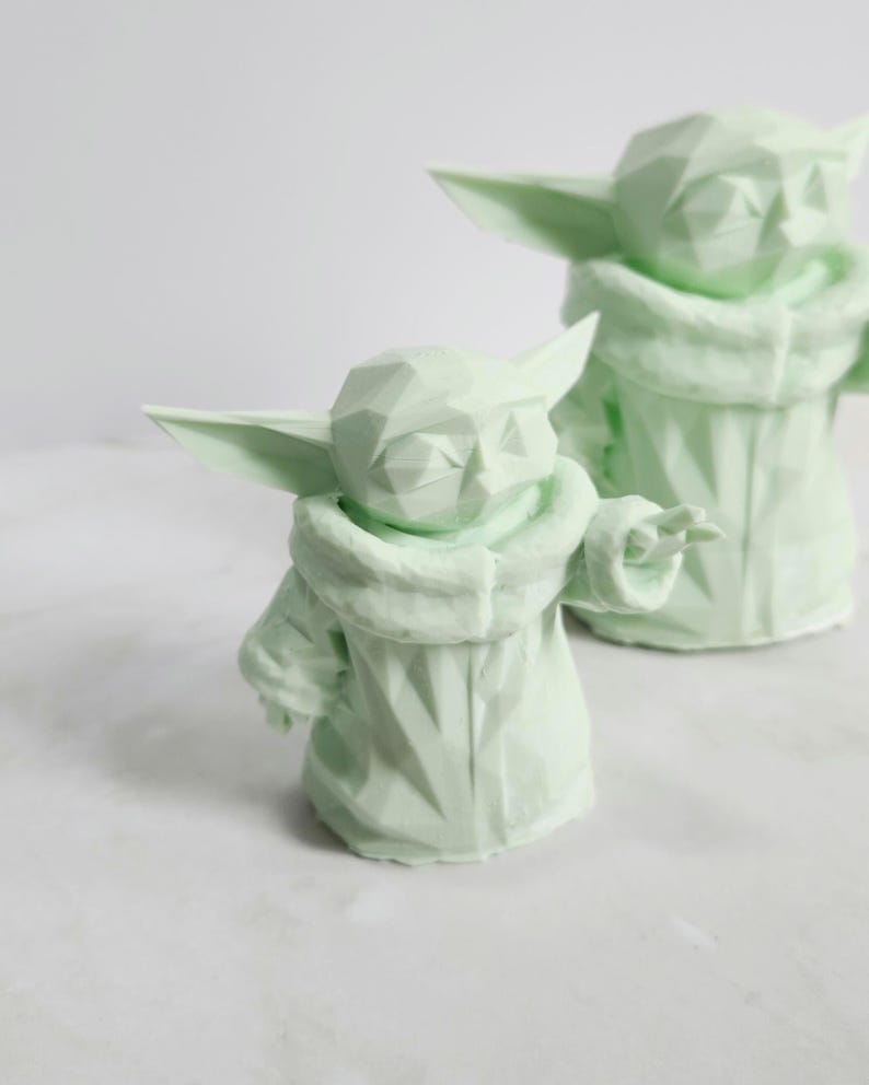 Pu&ograve; includere: Due sculture geometriche verde chiaro di un personaggio con grandi orecchie e un mantello con cappuccio. Le sculture hanno un design sfaccettato, che conferisce loro un aspetto moderno e angolare. Una scultura &egrave; in primo piano, con un'altra leggermente sfocata sullo sfondo.