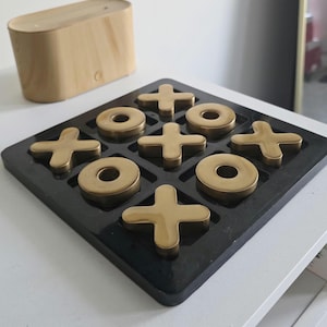 May include: A black tic-tac-toe board with gold-coloured X's and O's. A wooden, rectangular box is in the background. The game board is on a white surface.