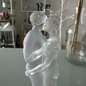 Könnte beinhalten: Eine klare Glasskulptur, die zwei Figuren in einer Umarmung zeigt, wobei eine Figur die andere küsst. Die Skulptur ist detailliert und zeigt die Formen der Figuren. Der Hintergrund ist verschwommen, mit einer Glasflasche und dekorativen Elementen.