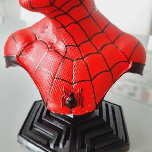 May include: A red and black Spider-Man bust figurine. The bust features a textured red surface with black web detailing and a black spider emblem on the chest. The figurine sits on a black geometric base.