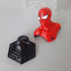May include: A red and black Spider-Man bust with a black base. The bust features the character's iconic mask and costume design. The base is a geometric shape with a textured surface. The bust is a collectible item.
