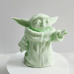 Pu&ograve; includere: Scultura geometrica verde chiaro di un personaggio con grandi orecchie e una veste. La figura &egrave; in posa con un braccio teso e la superficie ha un design sfaccettato. La scultura &egrave; su una superficie bianca.