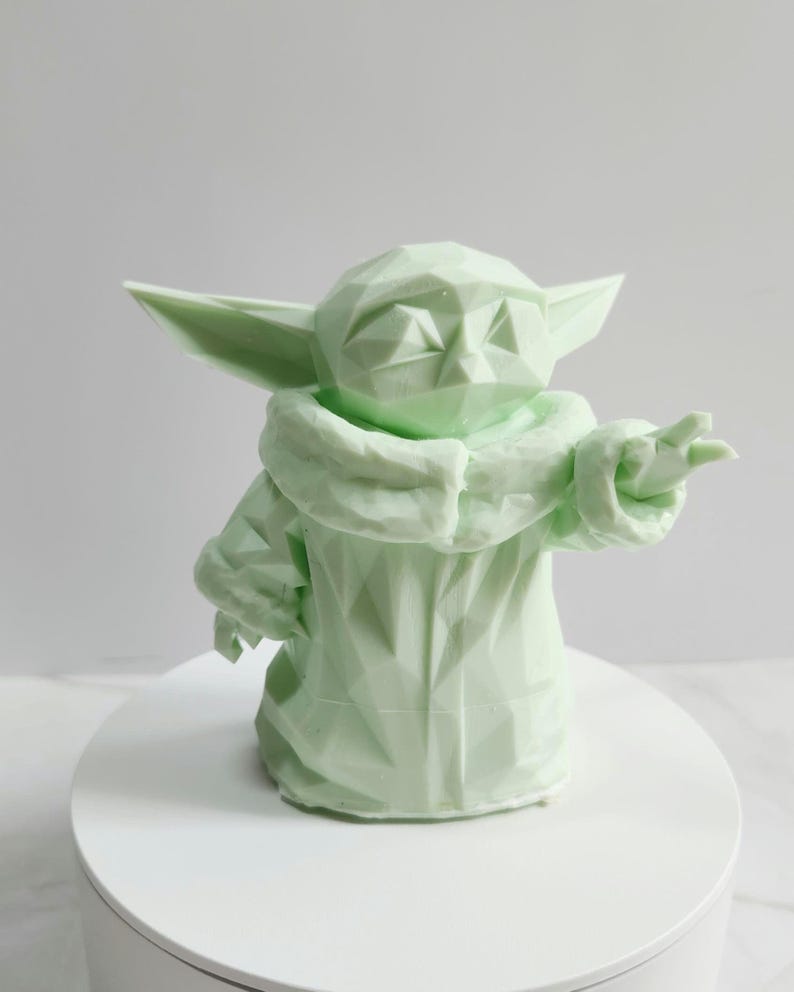 Pu&ograve; includere: Scultura geometrica verde chiaro di un personaggio con grandi orecchie e una veste. La figura &egrave; in posa con un braccio teso e la superficie ha un design sfaccettato. La scultura &egrave; su una superficie bianca.