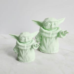 Pu&ograve; includere: Due sculture geometriche verde chiaro di un personaggio con grandi orecchie e un cappotto con cappuccio. Le sculture sono in posa eretta con un braccio teso. Le figure sono su una superficie bianca.