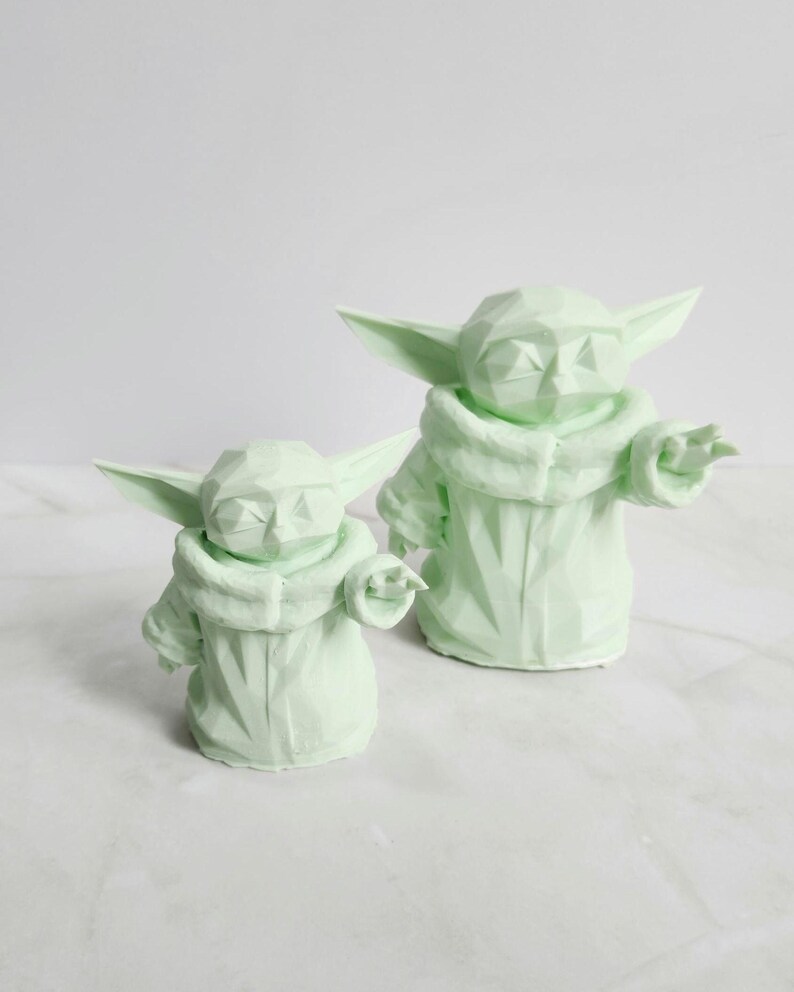 Pu&ograve; includere: Due sculture geometriche verde chiaro di un personaggio con grandi orecchie e un cappotto con cappuccio. Le sculture sono in posa eretta con un braccio teso. Le figure sono su una superficie bianca.
