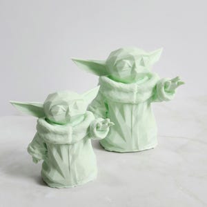 Pu&ograve; includere: Due sculture geometriche verde chiaro di un personaggio con grandi orecchie e un cappotto con cappuccio. Le sculture sono in un design stilizzato, low-poly, con sfaccettature visibili. Una scultura &egrave; leggermente pi&ugrave; piccola dell'altra.