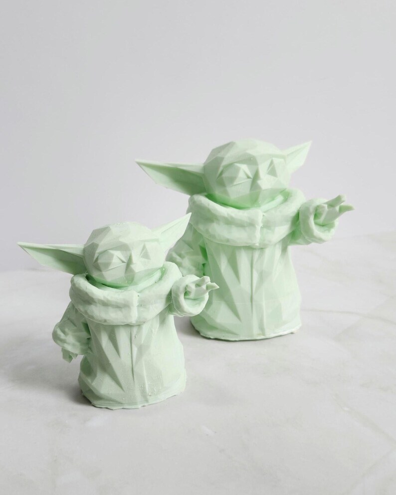 Pu&ograve; includere: Due sculture geometriche verde chiaro di un personaggio con grandi orecchie e un cappotto con cappuccio. Le sculture sono in un design stilizzato, low-poly, con sfaccettature visibili. Una scultura &egrave; leggermente pi&ugrave; piccola dell'altra.