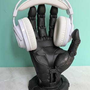 May include: White over-ear headphones sit on a black robotic hand-shaped stand. The headphones have white earcups and a padded headband. The stand is detailed with mechanical features and sits on a round base. The background is a light teal colour.