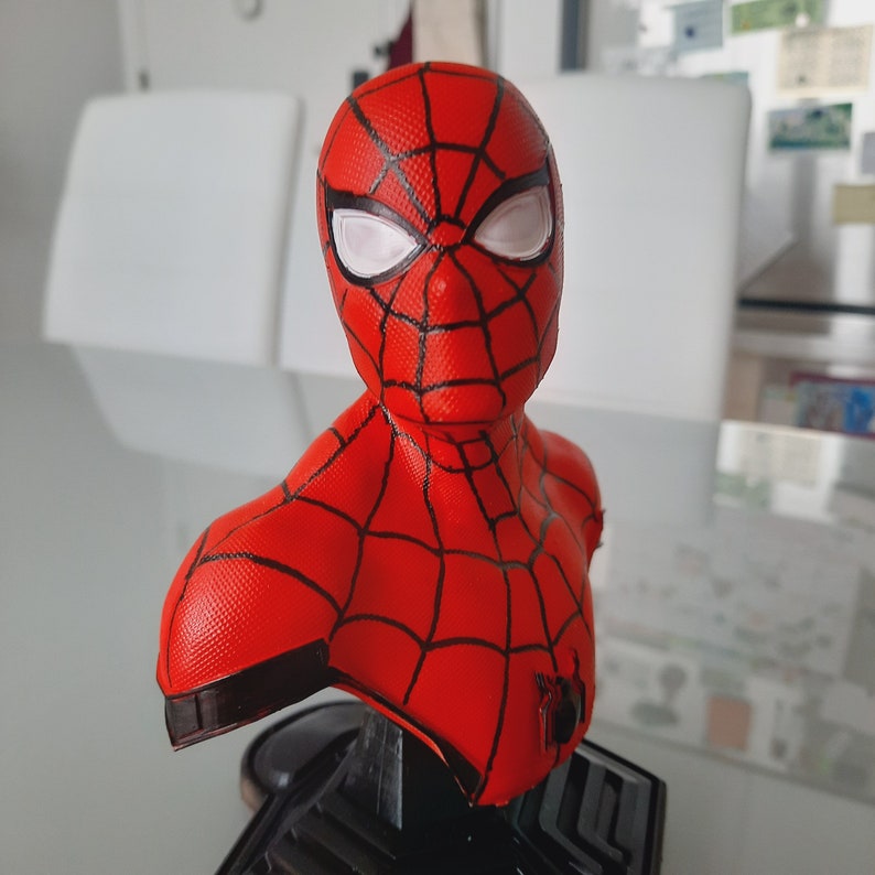 May include: A red and black Spider-Man bust figurine. The bust features the character's mask with white eye lenses and a webbed pattern. The figurine is mounted on a black base.