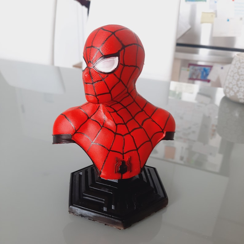 May include: A red and black Spider-Man bust figurine. The bust features the character's mask with white eye lenses and a web pattern. The figurine is mounted on a black, geometric base.