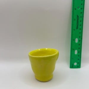 May include: A bright yellow ceramic cup with a smooth, glossy finish and a slightly flared rim. A green plastic Trailmaker ruler is positioned vertically next to the cup, showing the cup stands approximately 2.5 inches in height.