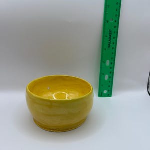 May include: A bright yellow ceramic bowl with a smooth, glossy finish and a rounded shape. A green plastic ruler is positioned vertically next to the bowl, showing the bowl stands approximately 2.5 inches in height.