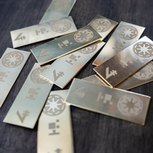 Può includere: Una collezione di targhette rettangolari metalliche con disegni incisi. Ogni targhetta presenta un emblema circolare, un simbolo stilizzato e altre forme geometriche. Le targhette sono sparse su una superficie di legno scuro, creando una disposizione visivamente accattivante.
