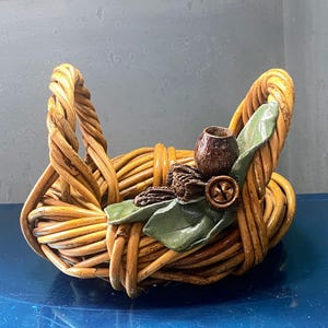 May include: A woven basket with two arched handles. The basket is made of light brown, twisted material. It is decorated with a green leaf, a small brown pot, and a wooden button.