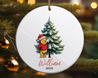 Può includere: Ornamento natalizio rotondo e bianco con un'illustrazione di Winnie the Pooh con cappello da Babbo Natale e maglione rosso, accanto a un albero di Natale verde. Il nome "William" e l'anno "2025" sono scritti in rosso.