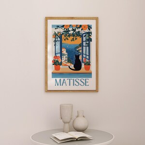 以下が含まれることがあります： 海岸の景色を望む窓辺に黒猫のシルエットが描かれた額入りのアートプリント。プリントには青色の「MATISSE」の文字、オレンジ色の花、鉢植えの植物が含まれています。フレームは木製です。