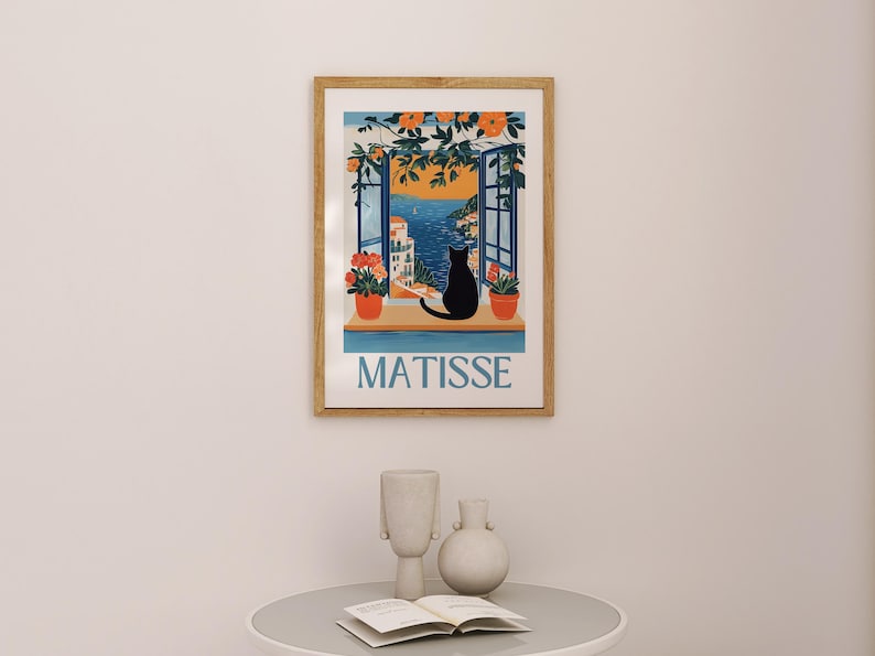以下が含まれることがあります： 海岸の景色を望む窓辺に黒猫のシルエットが描かれた額入りのアートプリント。プリントには青色の「MATISSE」の文字、オレンジ色の花、鉢植えの植物が含まれています。フレームは木製です。