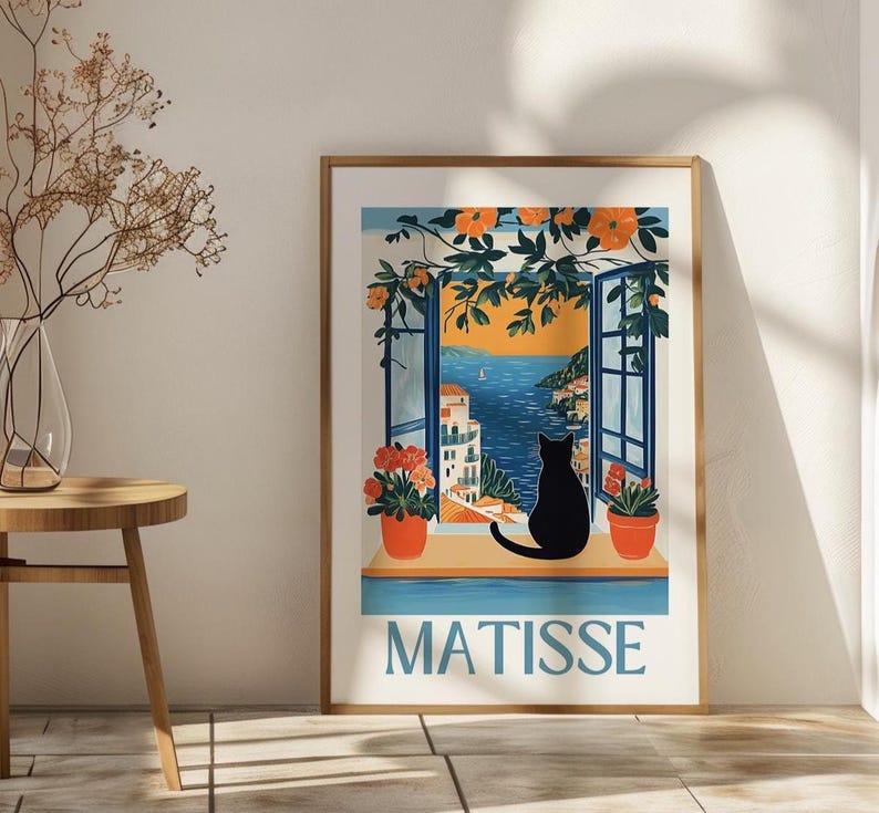以下が含まれることがあります： 海岸の風景を眺める開いた窓から外を眺める黒猫のシルエットが描かれた、額入りのアートプリントです。「MATISSE」の文字が薄い青色でプリントされています。オレンジ色の花と緑が窓を縁取っています。