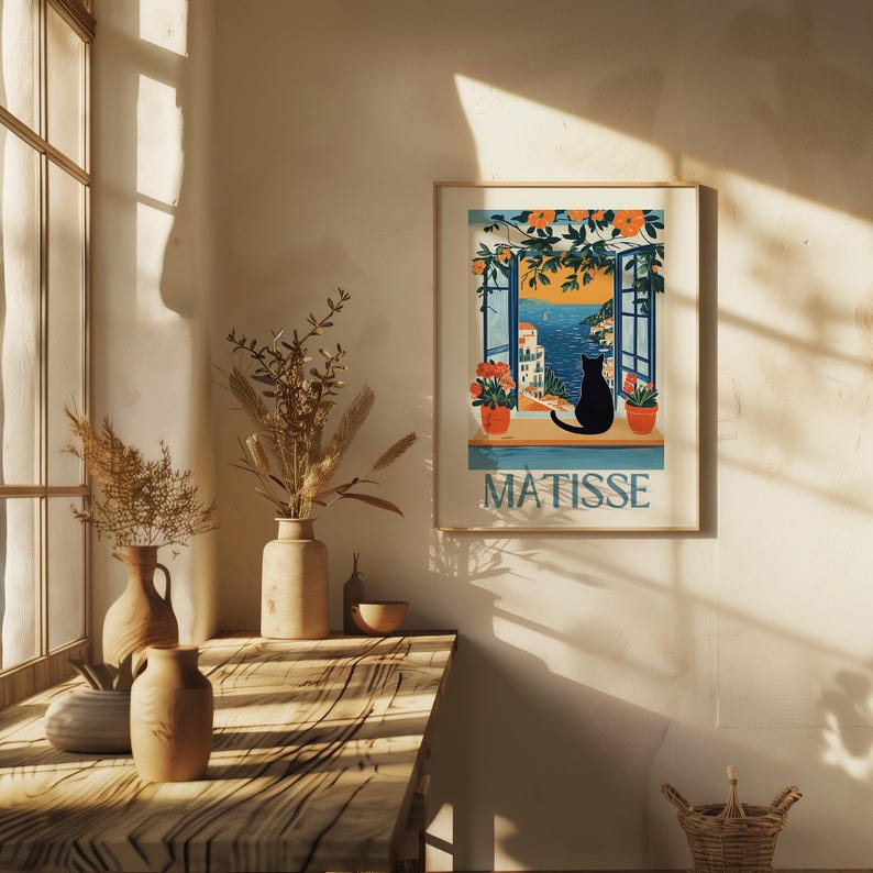 以下が含まれることがあります： 海岸の風景を窓から眺める黒猫のシルエットが描かれた、額入りのアートプリント。プリントには、青いフォントで「MATISSE」の文字が含まれています。アートワークは金のフレームに入っており、クリーム色の壁に飾られています。