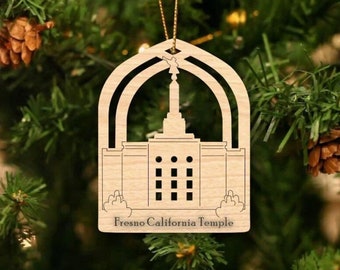 Puede incluir: Un adorno navideño de madera con forma del Templo de Fresno, California. El adorno está recortado con un diseño detallado del templo y tiene las palabras "Fresno California Temple" inscritas en la parte inferior.