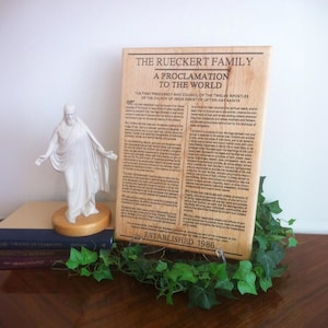 May include: A wooden plaque with the text "The Rueckert Family, A Proclamation to the World, Established 1986". The plaque is a light brown color and has a rustic appearance. The text is in a black font and is centered on the plaque.
