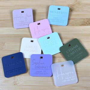 Puede incluir: Una colecci&oacute;n de etiquetas de nombre personalizadas en varios colores pastel, incluyendo lavanda, rosa y azul. Cada etiqueta presenta un nombre, informaci&oacute;n de contacto y un peque&ntilde;o agujero para adjuntar. Los nombres est&aacute;n grabados en una fuente elegante.