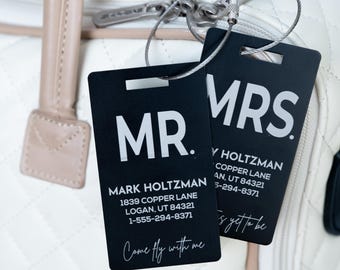 May include: Two rectangular black luggage tags with white text attached to a quilted white bag. One tag reads MR. MARK HOLTZMAN 1839 COPPER LANE LOGAN, UT 84321 1-555-294-8371 Come fly with me. The other reads MRS. Y HOLTZMAN 1839 COPPER LANE LOGAN, UT 84321 1-555-294-8371.