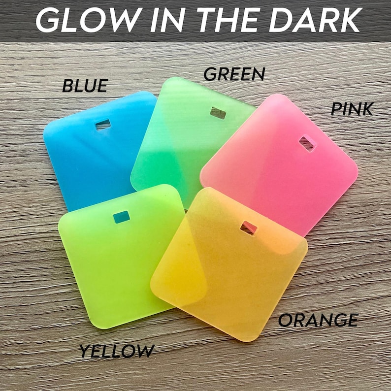 Puede incluir: Colecci&oacute;n de cinco accesorios cuadrados de silicona que brillan en la oscuridad en azul, verde, rosa, amarillo y naranja. Cada uno tiene un peque&ntilde;o agujero rectangular. El texto "GLOW IN THE DARK" est&aacute; en la parte superior.