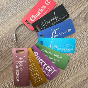 May include: A set of seven rectangular metal luggage tags in red, black, blue, teal, green, purple, and gold, all attached to a silver wire cable. Each tag features custom printed names and phone numbers, including Charles G., Hannah Anderson, jr, and Mylie Rueckert.