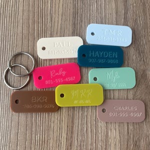 May include: A collection of eight rectangular acrylic key tags in various colors including white, light blue, dark blue, hot pink, mint green, brown, lime green, and mauve. Each tag features engraved names and phone numbers with two metal rings.