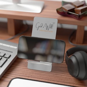 May include: A white card with the text "God's Will is what I want for my life" in black cursive writing with a small heart symbol. The card is propped up on a black smartphone stand on a wooden desk. A black pair of headphones are on the desk.