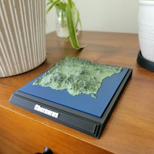 Puede incluir: Un modelo de mapa topográfico en 3D de Chernarus que presenta un relieve de paisaje verde texturizado montado sobre una base cuadrada azul plana. La base está dentro de un marco negro con el texto blanco Chernarus impreso en el borde frontal.