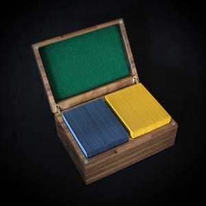 May include: A wooden playing card box with a green felt lining. Inside are two decks of cards, one blue and one yellow. The box is crafted from dark wood, showcasing a natural grain pattern. The box is open.