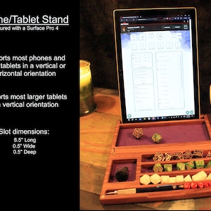 May include: A wooden phone and tablet stand with a purple felt lining. The stand has a slot for holding a tablet or phone in a vertical orientation. The stand also has a compartment for dice and other gaming accessories. The stand is pictured with a Surface Pro 4 tablet.