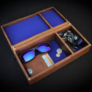 Könnte beinhalten: Eine Holz-Herrenbox mit mehreren Fächern, ausgekleidet mit blauem Stoff. Die Box enthält Sonnenbrillen, eine Geldbörse, Münzen und eine gemusterte Krawatte. Die Box ist aus dunklem Holz.
