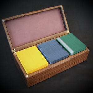 Könnte beinhalten: Eine Holzbox mit Scharnierdeckel, die drei Stapel Karten in Gelb, Blau und Grün enthält. Die Box besteht aus dunklem Holz mit rosa Innenraum und goldfarbenen Scharnieren.