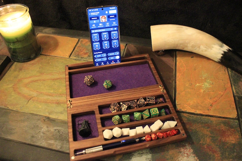 May include: A wooden box with a purple felt interior and compartments for dice and other gaming accessories. The box has a phone stand and a pen holder. There are several dice, some with metal accents, and a small metal figure in the box. The box is open and the phone is in the stand.
