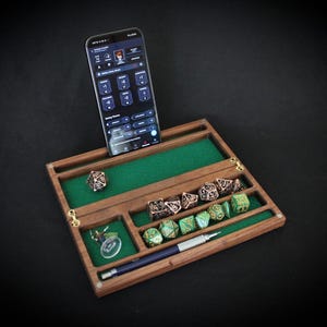 May include: A wooden dice rolling tray with a green felt interior, holding various polyhedral dice in bronze and green. A smartphone displaying an app is propped up in the tray. A pen and a keychain are also present.